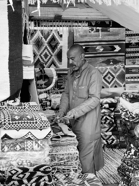 A man in a work uniform inspecting or organizing a display of woven rugs and textiles at a marketplace or shop. The rugs feature geometric patterns and are stacked on shelves and tables made of wood. 