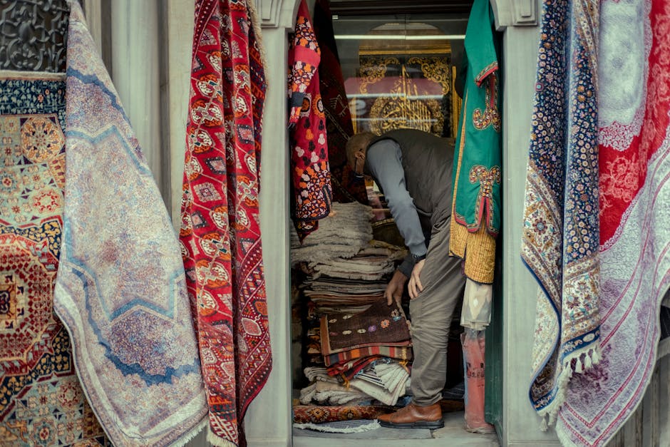 A man in a work uniform inspecting or organizing a display of woven rugs and textiles at a marketplace or shop. The rugs feature geometric patterns and are stacked on shelves and tables made of wood. 